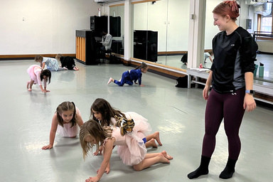 Young dancers crawl in a dance studio watched by their instructor. One dancer has a stuffed animal tiger on her back.