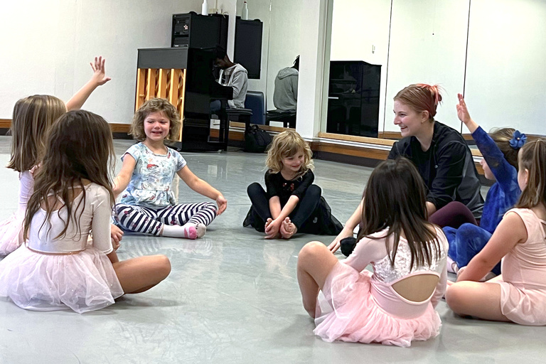 Young dancers sit in a circle with hands raised for their smiling teacher