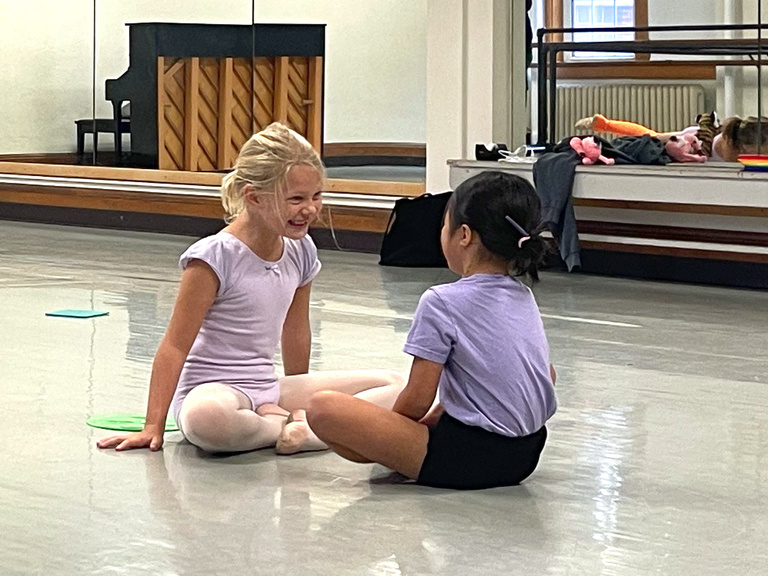 Two students sit on a dance studio floor smiling at each other. In the distance are colorful toys and a piano. 