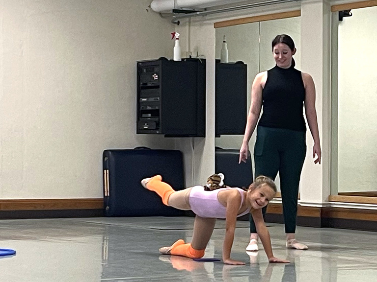A young ballet dancer has a big smile while she crawls. Her teacher watches nearby.